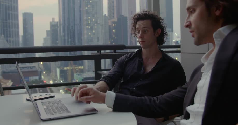 Two men work on a laptop while sitting at a table on an outdoor balcony