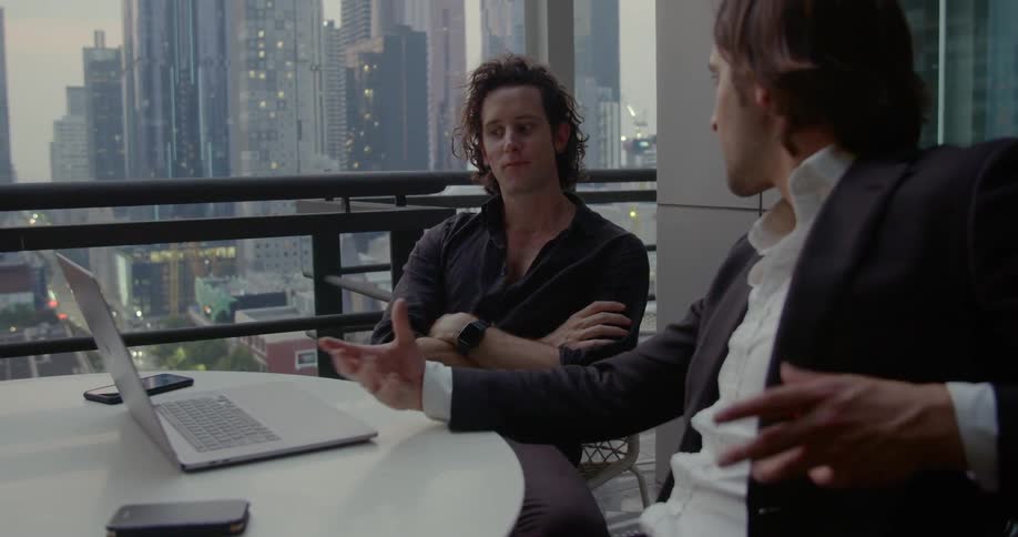 Two men work on a laptop while sitting at a table on an deck in a high rise building