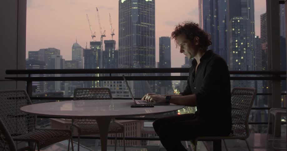 A man types on a laptop while sitting in front of a window with skyscrapers in the distance