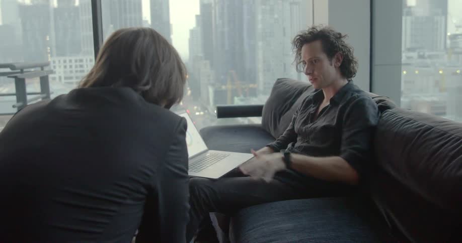 Two men sit in living room collaborating over laptop near large windows
