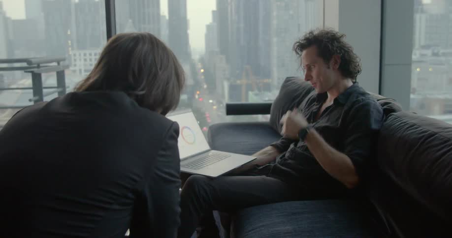 Two people in a room discuss a chart on a laptop screen