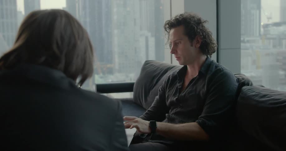 A man holds a laptop and uses the trackpad while sitting on a couch in an office talking to a colleague