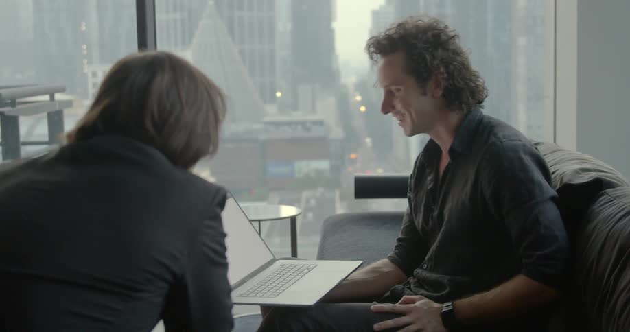 A man sits with his laptop on his knee and shows another man graphs on it