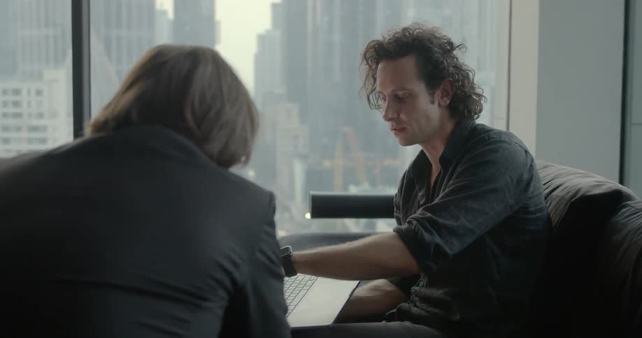 A man holds a laptop in his hand as he talks to a colleague while sitting on a sofa in an office