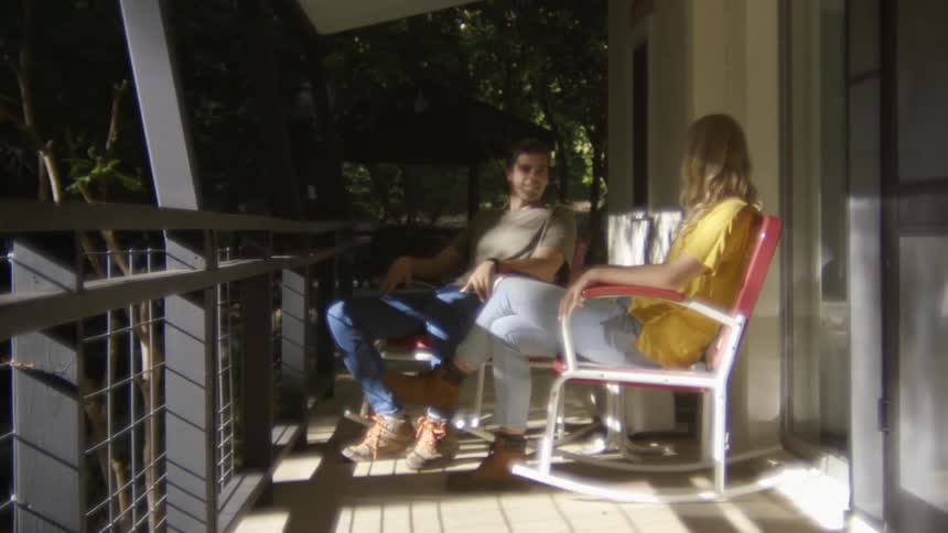 A man and woman sit on rocking chairs on a balcony surrounded by trees