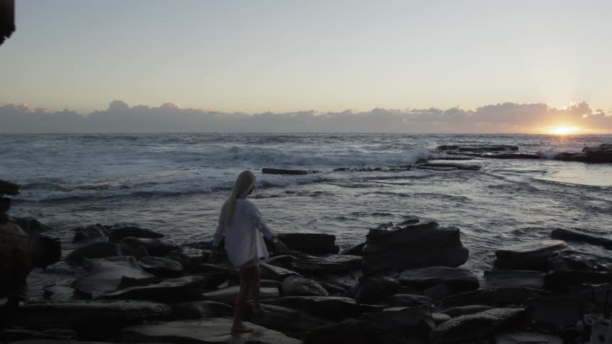A slow-motion clip of a woman walking on shoreline rocks during low tide at sunset
