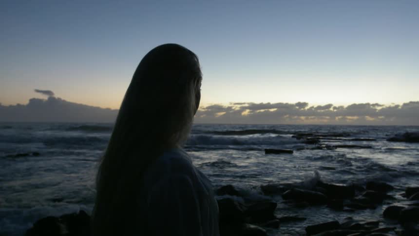A woman is standing by the water watching the sun set