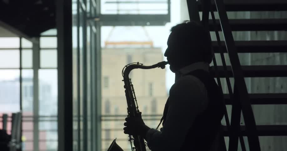 A man plays the saxophone standing next to a dark staircase