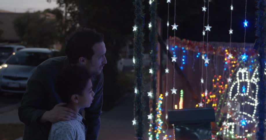 A dad hugs his son and points at a light display with hanging stars and colorful lights