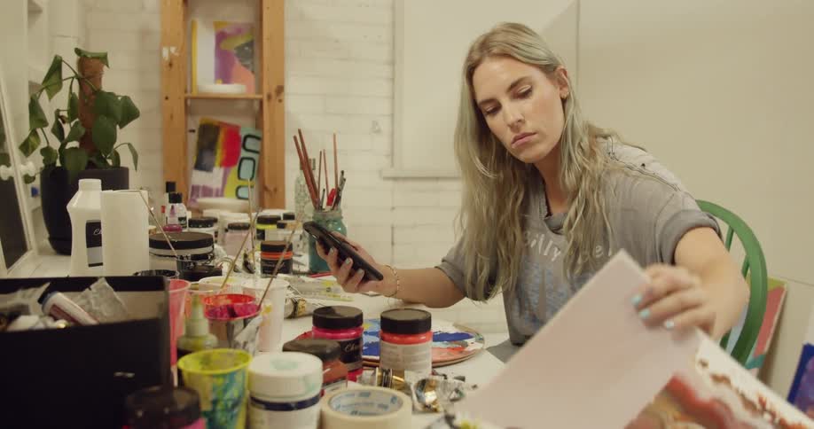A blonde woman sits at a table and looks at various paintings