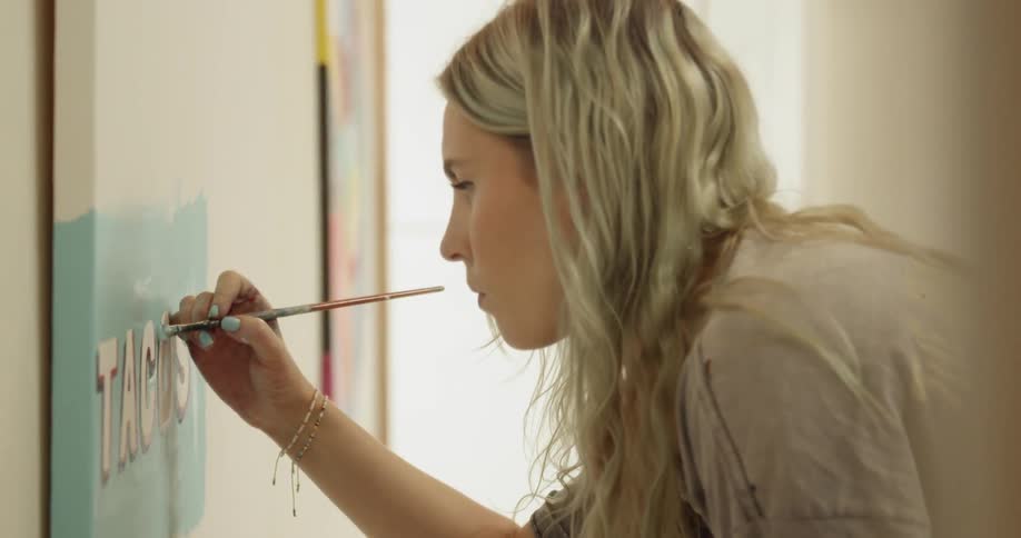 A woman uses a paintbrush to shade around letters painted on a canvas hanging on a wall