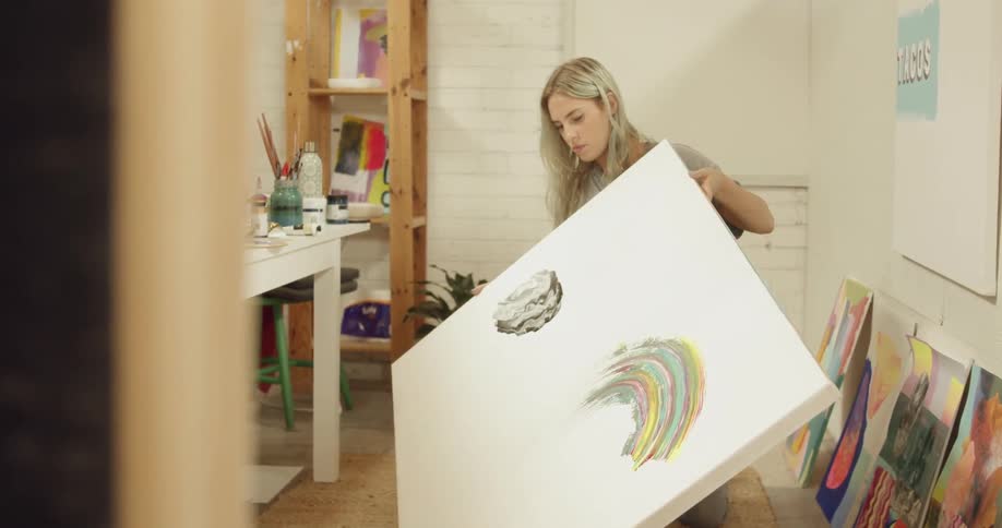 A woman holds up a large canvas with some paint on it in an art studio