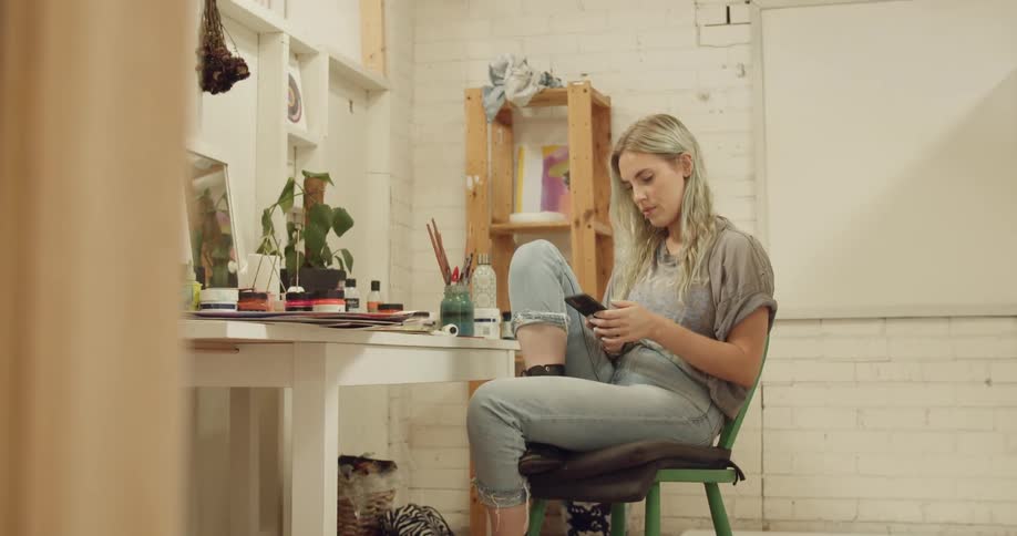A blonde woman sits on a green chair in a room while texting on her smartphone