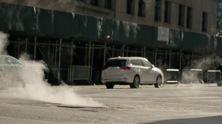 Steam billows from manhole covers on a street with cars parked near buildings