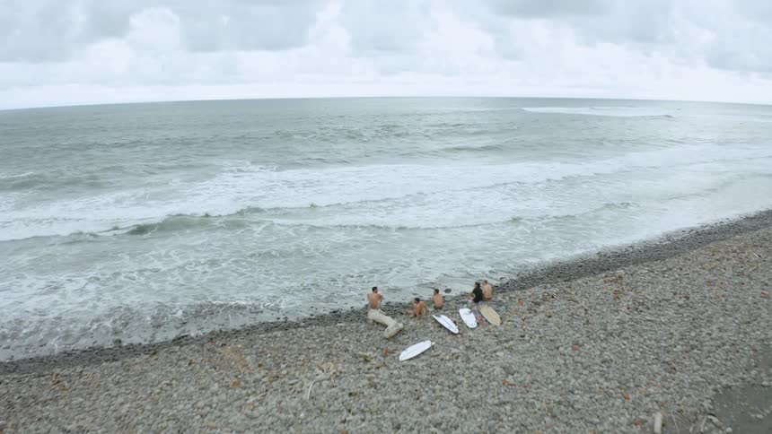 A group of surfers sit on a rocky coastline next to the ocean