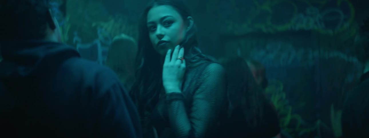 A young woman dances with a young man at a dark club with flashing lights and graffiti on the walls