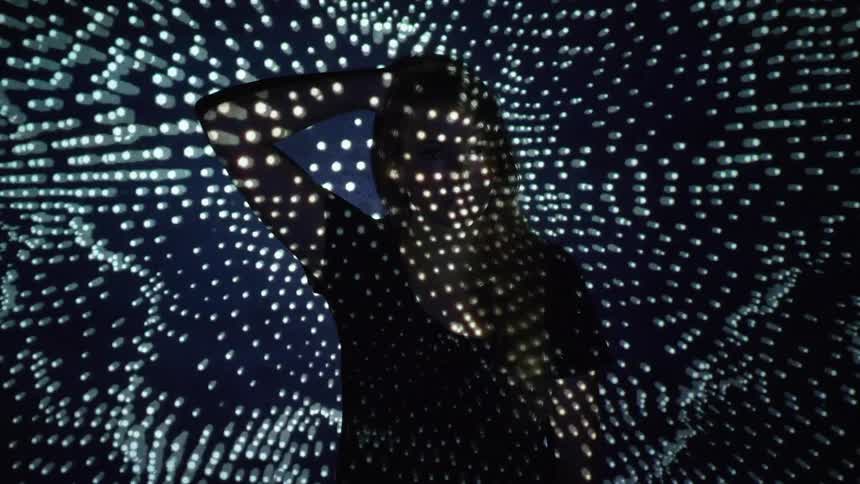 A girl stand and puts her hand behind her head as lights flash around her