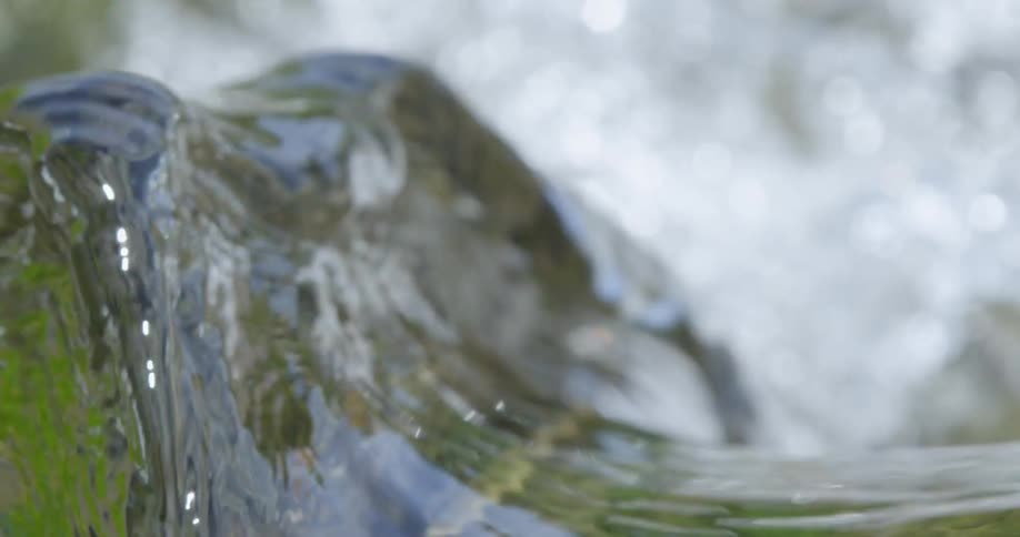 Clear water flowing in a river rises up and over a rock then splashes down below