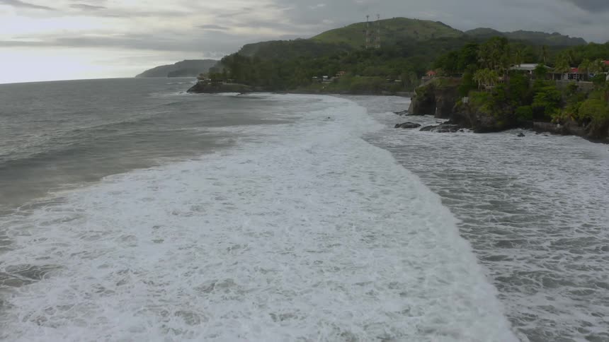 Ocean waves come to shore with palm trees and homes on the rocky coast