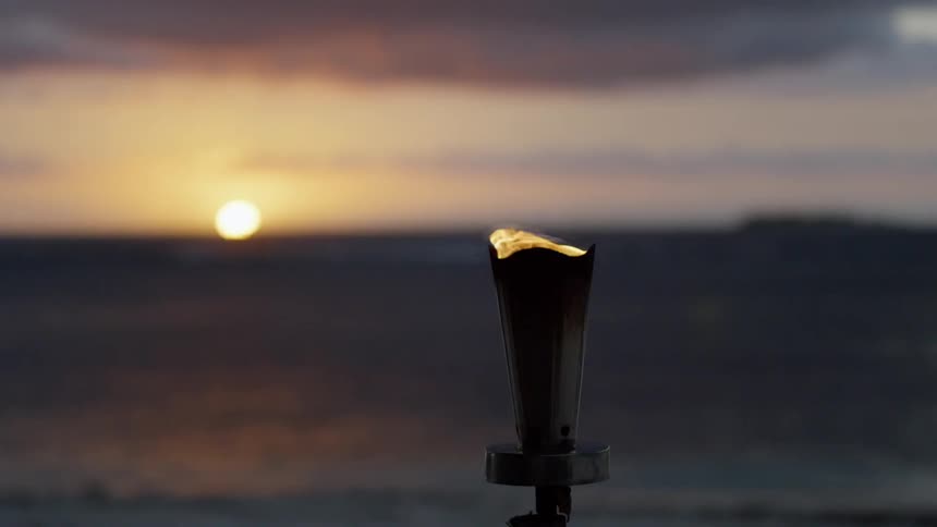A flame flickers atop a torch that is located near the beach as the sun is low in the sky in the background