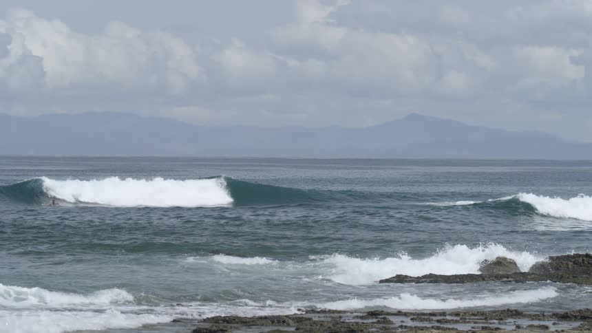 Surfer waits to catch wave under cloudy sky with mountains on the horizon