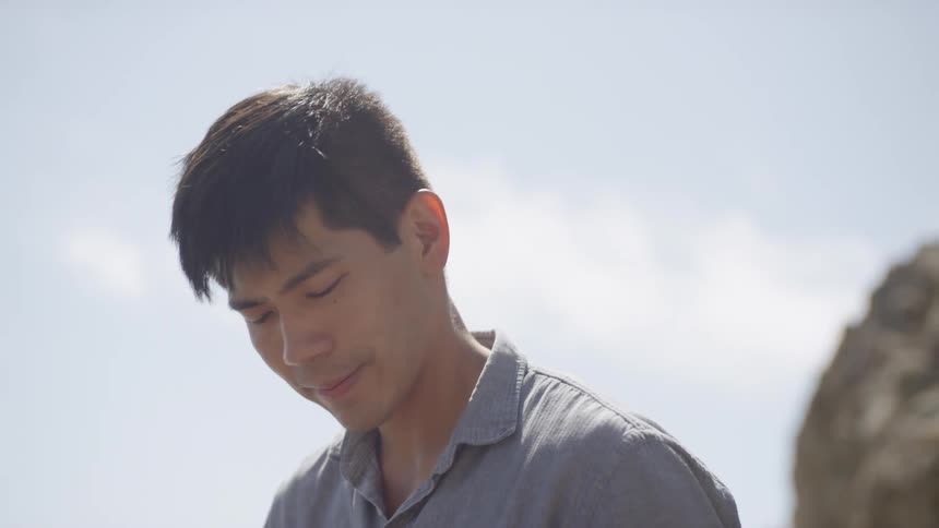 Asian man sitting down on a rock plays a ukulele next to the river