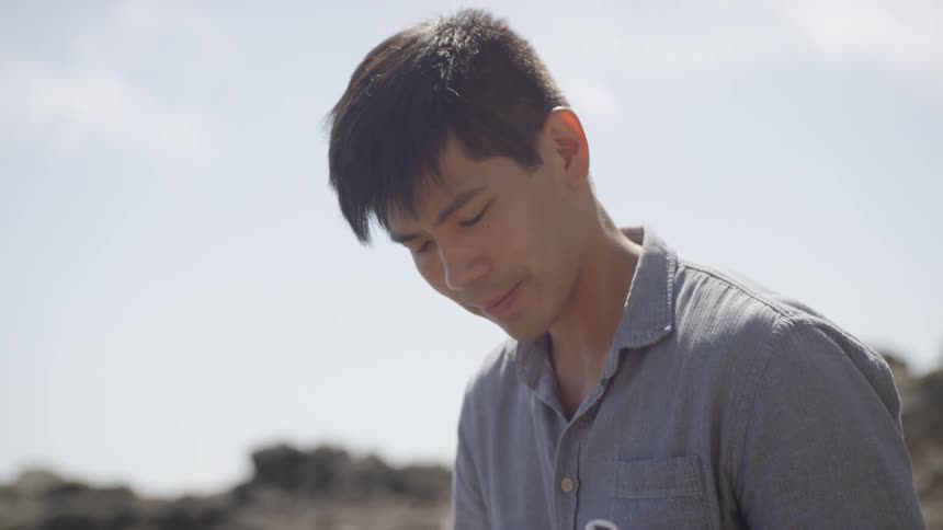 Asian man sitting on a rock plays on the ukulele next to the stream on a sunny day
