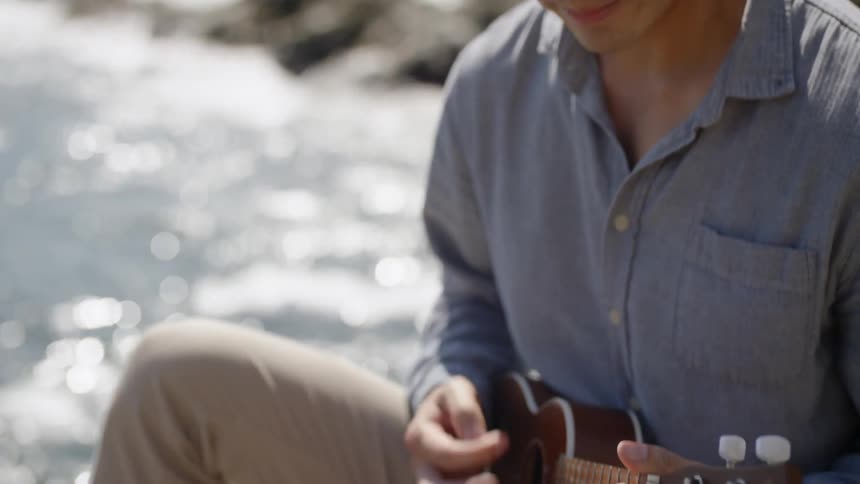 A man sits on a rocky coast line and plays a small guitar
