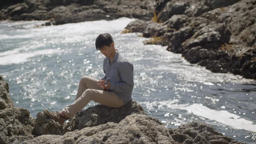 A young man sits atop a rock formation along the ocean shoreline where he strums on his ukulele