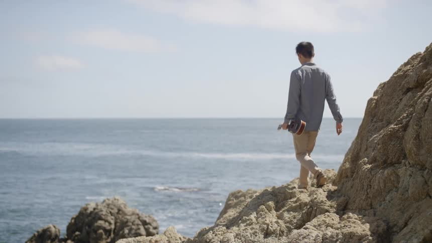 A man walks across a rocky beach while holding a ukulele