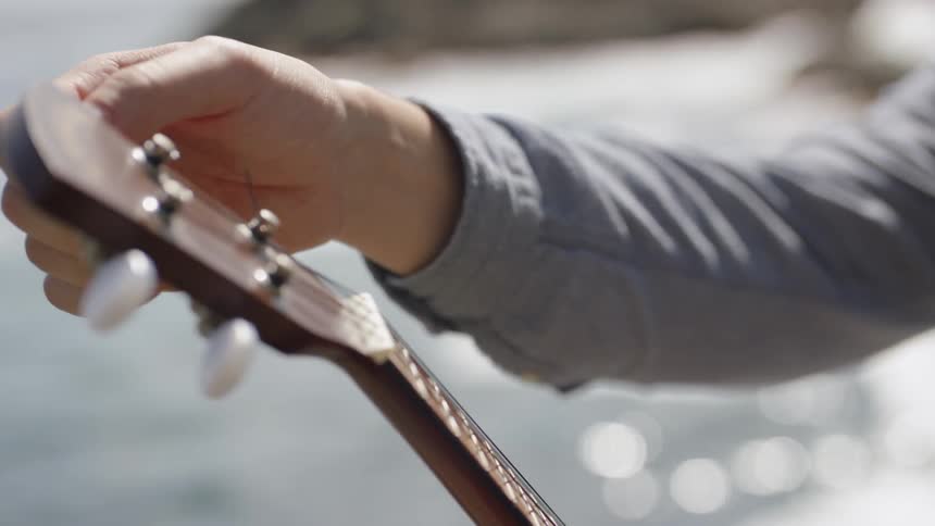 A man standing outside near a body of water on a sunny day tunes his acoustic guitar