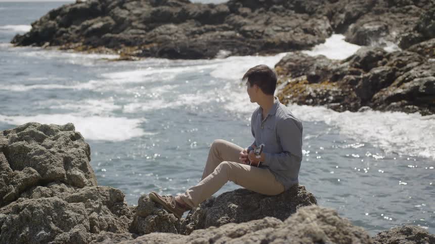 Asian man sitting on a rock plays on the ukulele by the sea