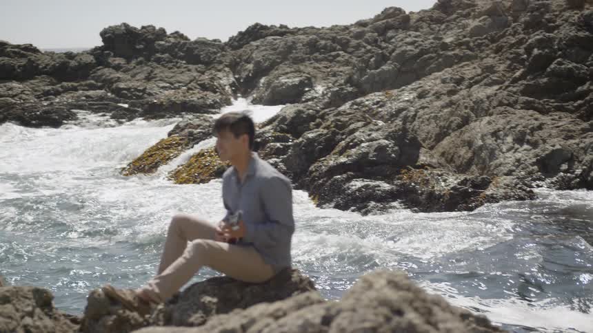 A young man sits on a rocky shore playing a guitar while ocean water swirls and splashes around him