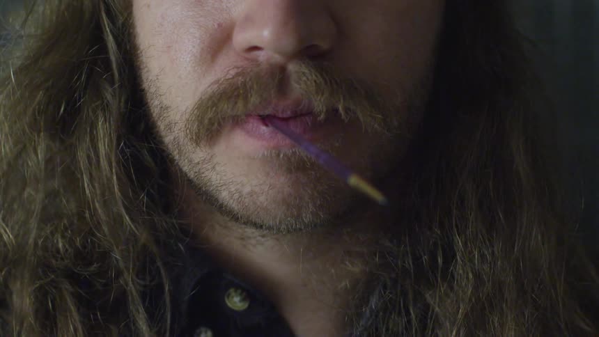 A scruffy man stands still with a small artist's paintbrush in his mouth