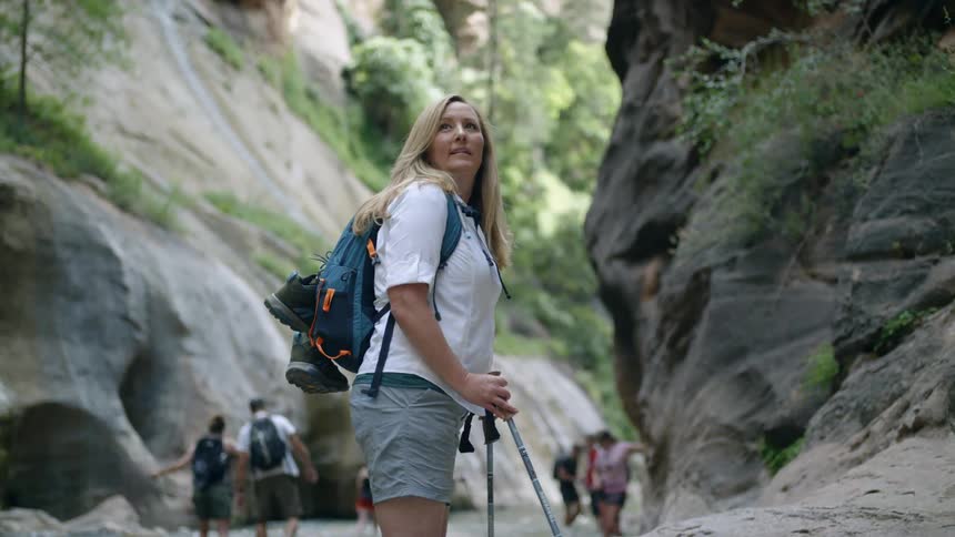A woman uses walking sticks though a shallow river and rocky terrain