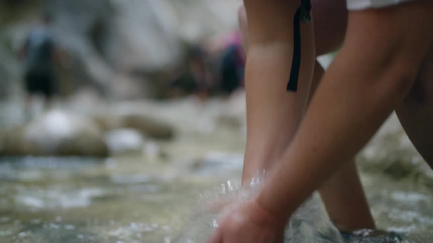 A woman standing in a stream reaches down into the water to wash her hands and arms