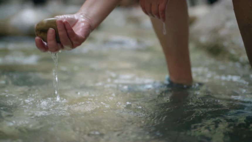 Person crouching in water picks up rock from river bed