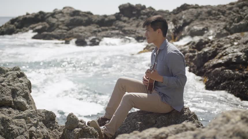 A young man sits on a rock by the ocean while strumming a guitar