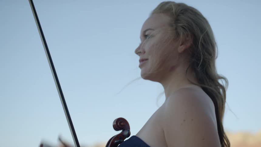 A woman holds a violin and bow outside in a rural terrain