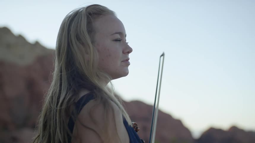 Blonde woman holding the violin tucks hair behind ear in the canyon