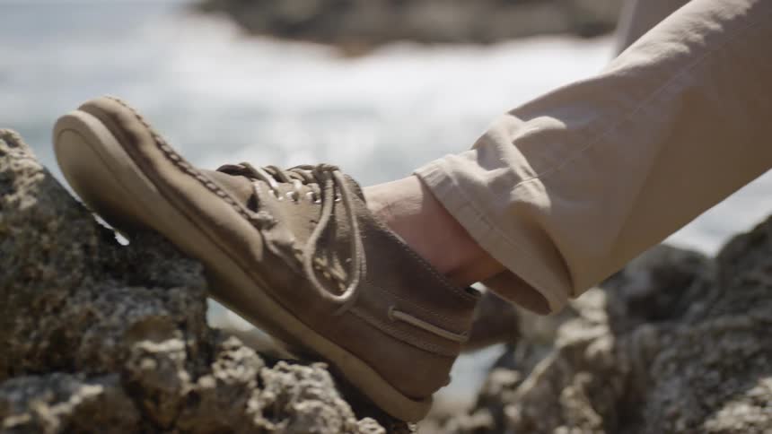 A man is playing ukulele and tapping his foot while sitting on some rocks near the ocean