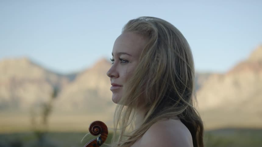Woman gazes at craggy lightly vegetated mountains while holding violin