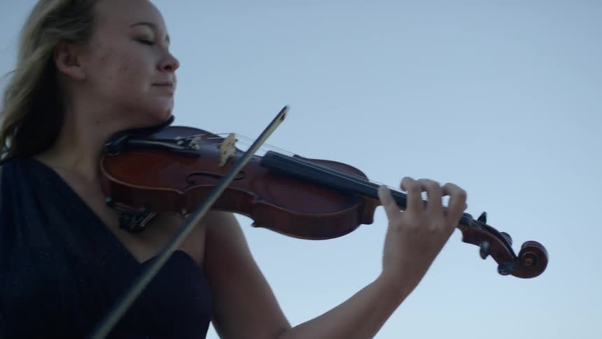 A blonde white female plays the violin outside under a clear blue sky