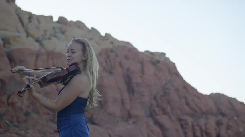 Blonde woman wearing a blue dress plays on the violin in the canyon