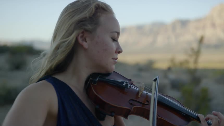 A white female violinist plays her instrument outdoors with mountains in the background