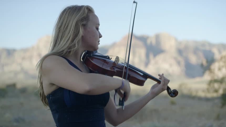An older blonde woman in a blue dress plays a violin outdoors