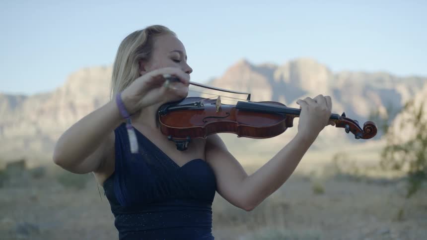 Blonde woman wearing a blue dress plays on the violin in the canyon