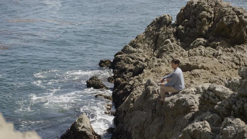 A young man with a musical instrument sits on a cliff looking out at the sea