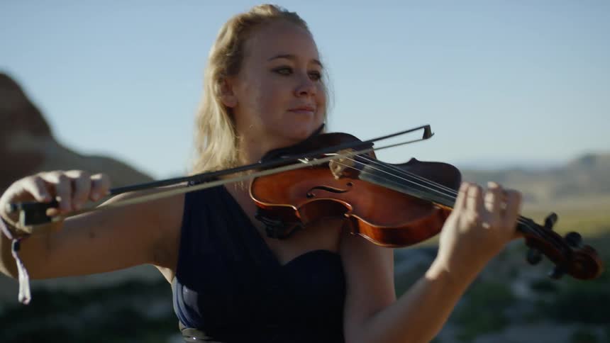 A woman wearing a dress plays a violin outdoors near a large rock formation