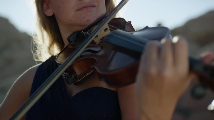 A woman in an evening gown stands outside in the sunlight while playing a violin